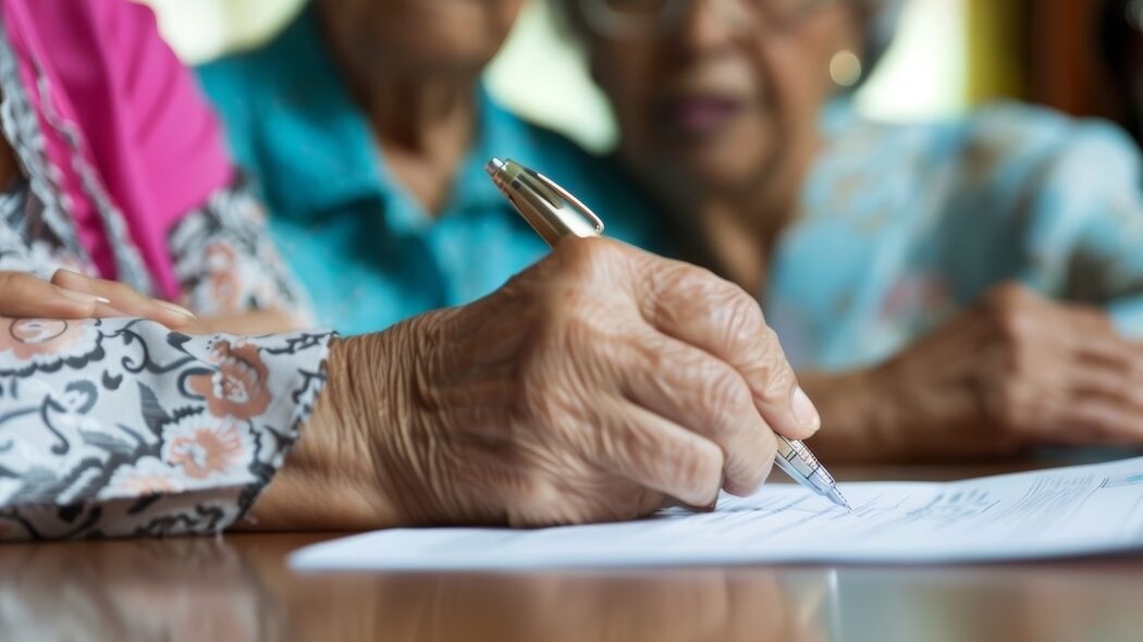 Elderly woman writing notes with a pen, with another senior woman in the background, highlighting the theme of companionship and shared experiences in later life.