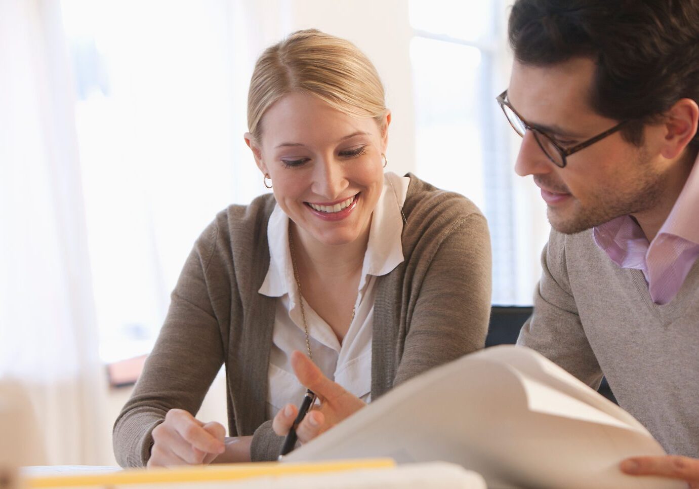 Two colleagues engaged in a discussion at a workspace, smiling and collaborating on a project with papers and a notebook in front of them.