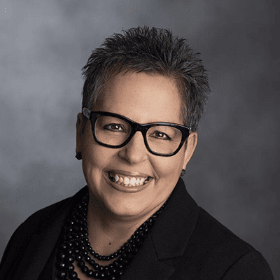 Professional headshot of a smiling woman with short, spiky gray hair and glasses, wearing a black jacket and necklace, set against a neutral gray background.