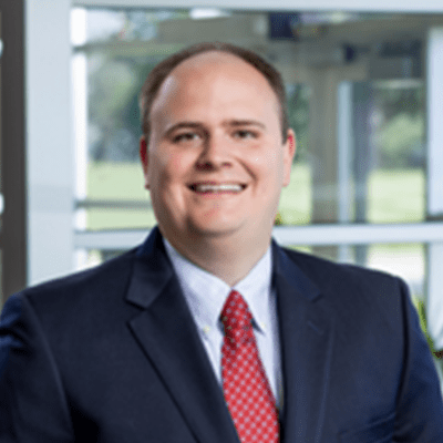 Professional portrait of a smiling man in a dark suit and red tie, standing in an office environment with large windows and greenery in the background.