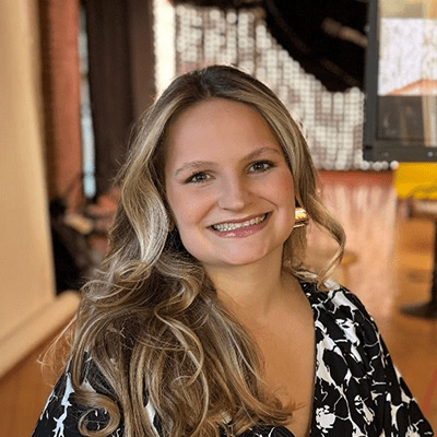 Smiling woman with long, wavy hair wearing a black and white patterned dress, standing indoors in a well-lit space with artistic decor in the background.